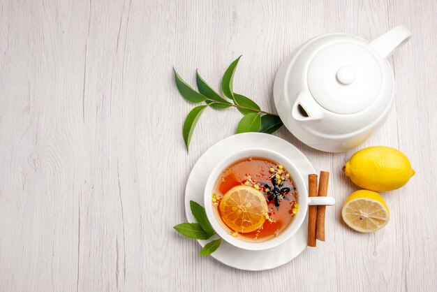 Vista superior de té y canela, una taza de té de hierbas, palitos de canela en el platillo, tetera de limón y hojas sobre la mesa blanca