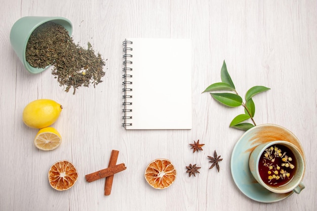 Vista superior de té de hierbas té de hierbas en la taza blanca junto al cuaderno blanco limón anís estrellado palitos de canela hierbas en la placa blanca