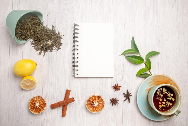 Vista superior de té de hierbas té de hierbas en la taza blanca junto al cuaderno blanco limón anís estrellado palitos de canela hierbas en la placa blanca