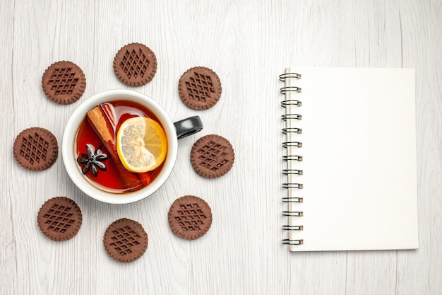 Vista superior de té de limón y canela redondeado con galletas y un cuaderno sobre la mesa de madera blanca