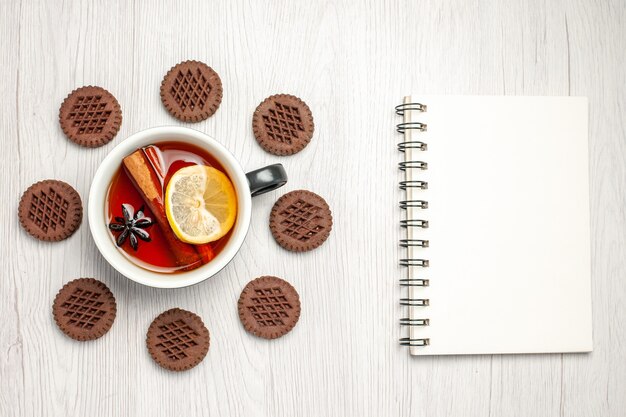 Vista superior de té de limón y canela redondeado con galletas y un cuaderno sobre la mesa de madera blanca