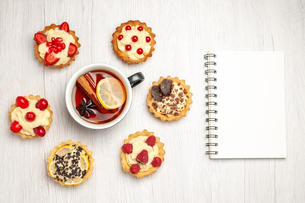 Vista superior de té de limón y canela redondeado con tartas y un cuaderno sobre la mesa de madera blanca