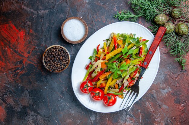 Vista superior tenedor de ensalada de verduras en placa ovalada diferentes especias en tazones pequeños en la mesa de color rojo oscuro