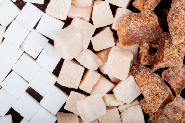 Vista superior de terrones de azúcar blanco y marrón y pedazos de azúcar de palma esparcidos sobre fondo de madera oscura