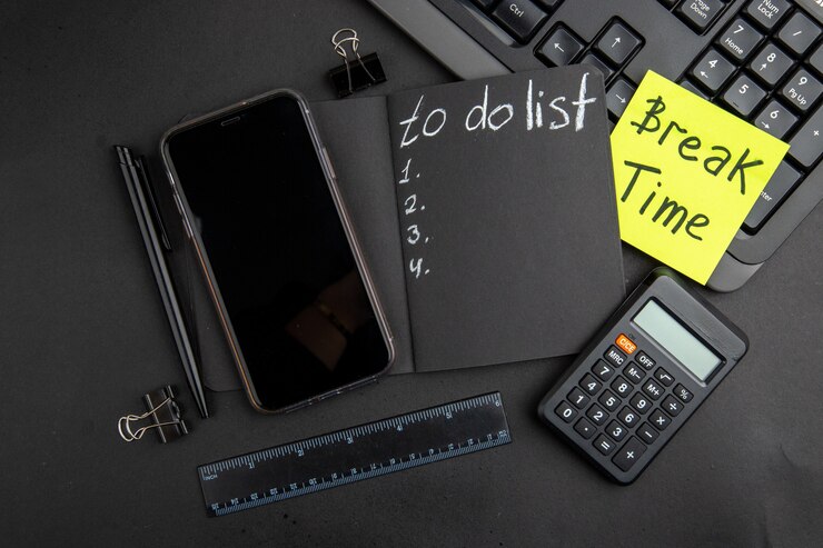 Foto gratuito vista superior del tiempo de descanso escrito en una nota adhesiva para hacer la lista en el bloc de notas negro, teléfono, lápiz, calculadora, regla, carpeta, clips, teclado, sobre tabla negra