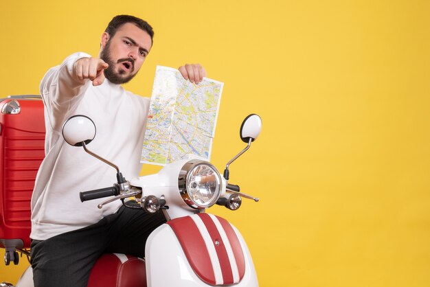 Vista superior del tipo curioso sentado en la motocicleta con la maleta sosteniendo el mapa apuntando hacia adelante sobre fondo amarillo aislado