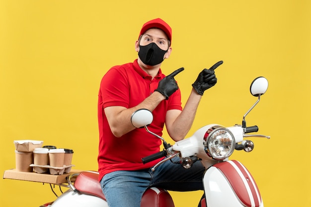Vista superior del tipo de mensajero curioso con blusa roja y guantes de sombrero en máscara médica entregando orden sentado en scooter apuntando algo