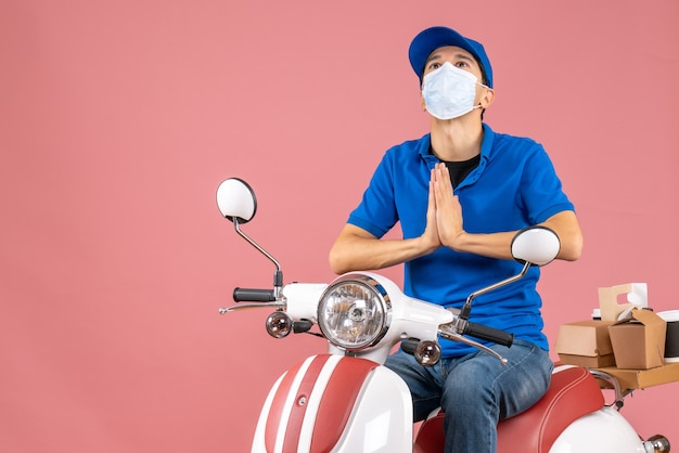 Vista superior del tipo de mensajero de ensueño con máscara médica con sombrero sentado en scooter entregando pedidos sobre fondo melocotón pastel