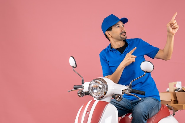 Foto gratuita vista superior del tipo de mensajero con sombrero sentado en scooter entregando pedidos devueltos apuntando hacia arriba sobre fondo melocotón pastel