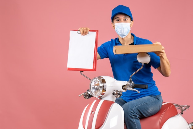 Vista superior del tipo de mensajero trabajador en máscara médica con sombrero sentado en scooter sosteniendo documento sobre fondo melocotón pastel