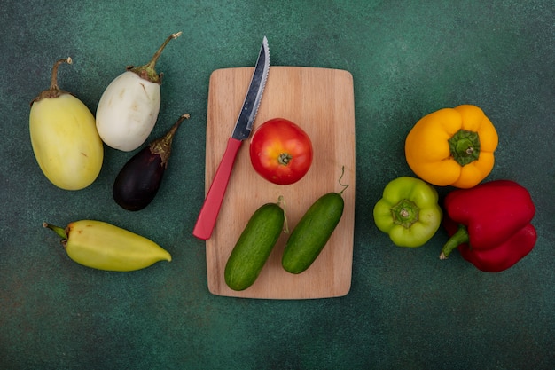 Vista superior de tomate con pepinos en una tabla de cortar con un cuchillo y pimientos de colores berenjenas sobre un fondo verde