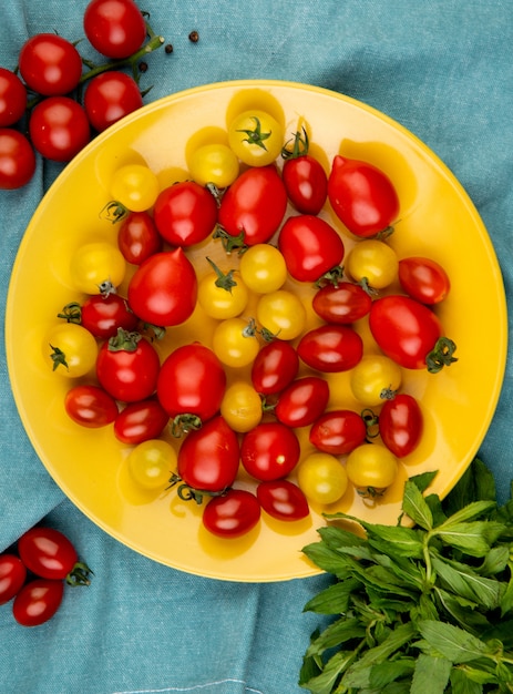 Vista superior de tomates amarillos y rojos en placa con hojas de menta verde en la mesa de tela azul