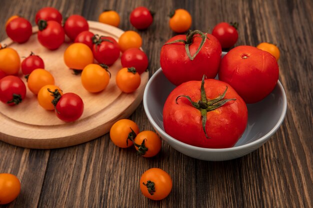 Vista superior de tomates cherry rojos y naranjas aislados en una tabla de cocina de madera con tomates de gran tamaño en un recipiente en una pared de madera