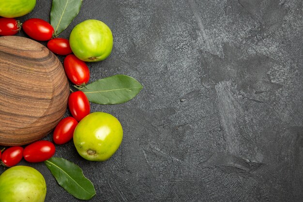 Vista superior de tomates cherry tomates verdes y hojas de laurel alrededor de una placa de madera sobre un suelo oscuro con espacio de copia