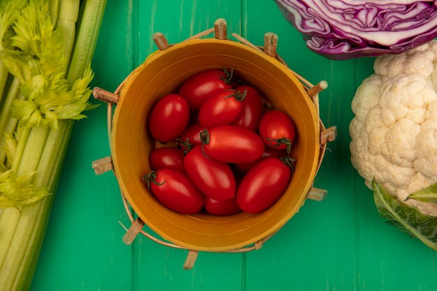 Vista superior de tomates ciruela roja en un balde con coliflor de repollo morado y apio aislado en una pared de madera verde