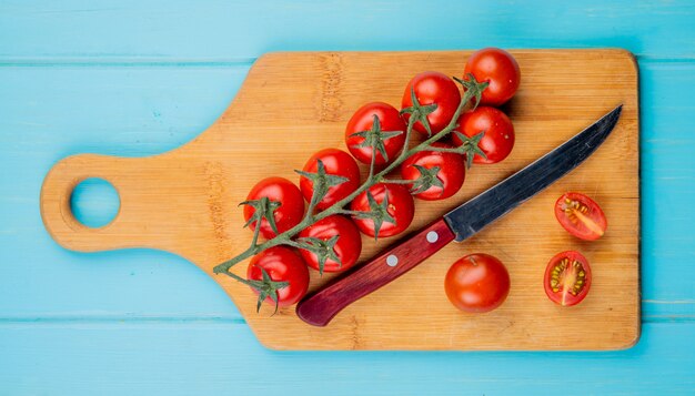 Vista superior de tomates cortados y enteros con cuchillo en la tabla de cortar sobre superficie azul