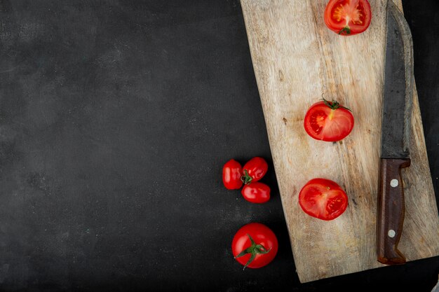 Vista superior de tomates frescos y mitades sobre tabla de cortar de madera junto a un cuchillo de cocina sobre fondo negro