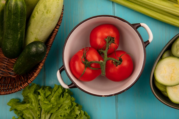 Vista superior de tomates orgánicos en un recipiente con calabacines y pepinos en un balde con lechuga y apio aislado sobre un fondo de madera azul