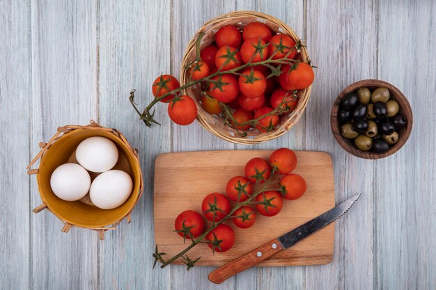 Vista superior de tomates en rama frescos en una tabla de cocina de madera con cuchillo con tomates en rama en un cubo y aceitunas en un cuenco de madera sobre un fondo de madera gris