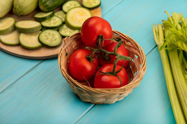 Vista superior de tomates rojos frescos en un balde con pepinos picados y calabacines en una tabla de cocina de madera con apio aislado en una superficie de madera azul