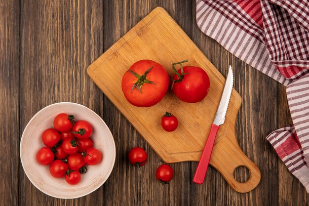 Vista superior de tomates rojos frescos en un recipiente con tomates en una tabla de cocina de madera con cuchillo sobre una superficie de madera