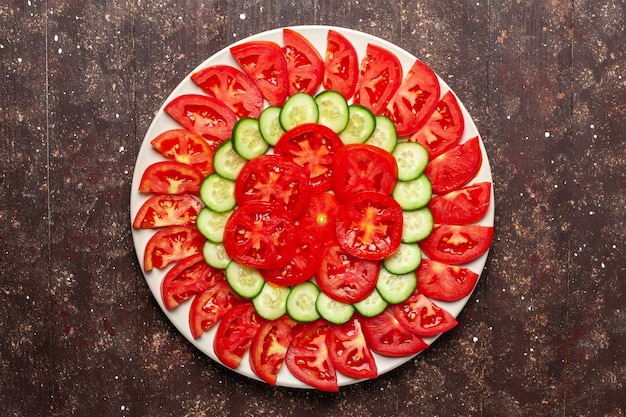 Vista superior de tomates rojos frescos en rodajas con ensalada fresca de pepinos en el espacio marrón