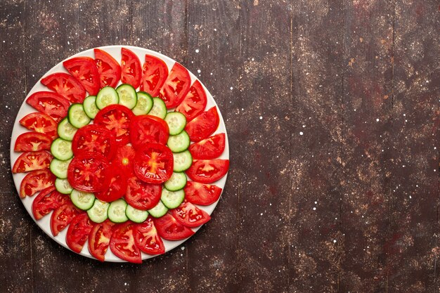 Vista superior de tomates rojos frescos en rodajas con ensalada fresca de pepinos en el espacio marrón