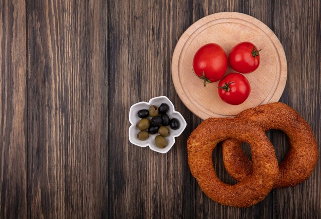 Vista superior de tomates rojos frescos en una tabla de cocina de madera con aceitunas en un recipiente y bagels aislado sobre un fondo de madera con espacio de copia