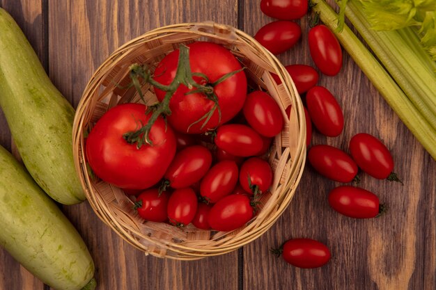 Vista superior de tomates rojos orgánicos en un balde con calabacines y apio aislado en una pared de madera