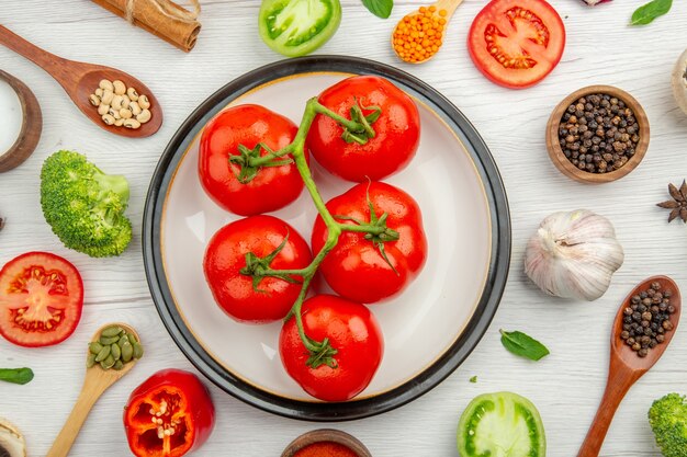 Vista superior de tomates rojos en un plato, pimienta negra en un tazón, cucharas de madera, ajo, brócoli en mesa gris