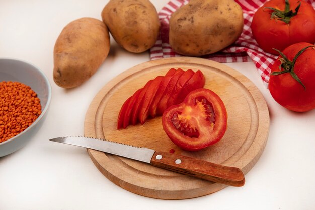 Vista superior de tomates rojos en rodajas en una tabla de cocina de madera con un cuchillo con lentejas naranjas en un recipiente con tomates enteros y papas aisladas en un paño marcado en una pared blanca