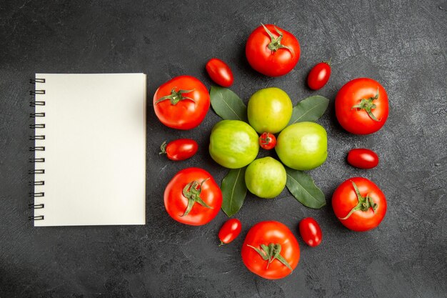 Vista superior de tomates rojos y verdes hojas de laurel alrededor de un tomate cherry y un cuaderno sobre suelo oscuro