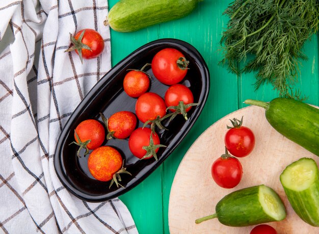 Vista superior de tomates en un tazón y pepino eneldo sobre tela escocesa y superficie verde