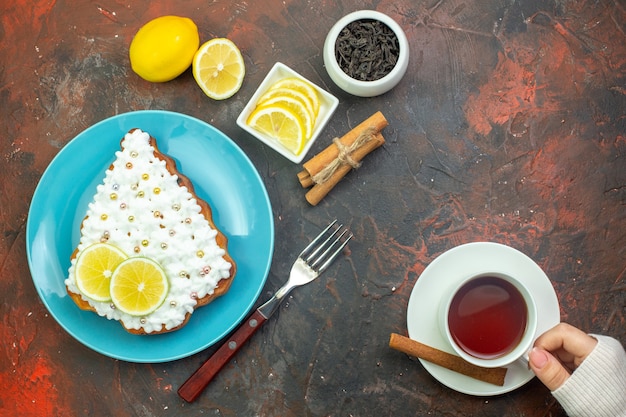 Vista superior de la torta con limón en placa azul rodajas de limón en un tazón tenedor taza de té en palitos de canela de mano femenina sobre fondo rojo oscuro