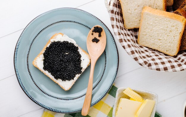Vista superior tostadas con caviar negro con una cuchara en un plato con mantequilla y pan en una cesta