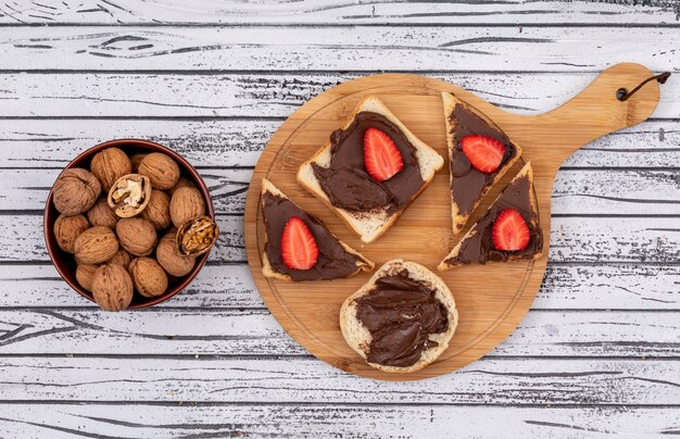 Vista superior de tostadas de desayuno con chocolate y fresa en tabla de cortar y nueces en un tazón sobre una superficie de madera blanca horizontal