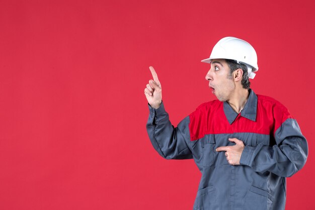 Vista superior del trabajador joven confundido en uniforme con casco apuntando hacia arriba y el lado derecho en la pared roja aislada