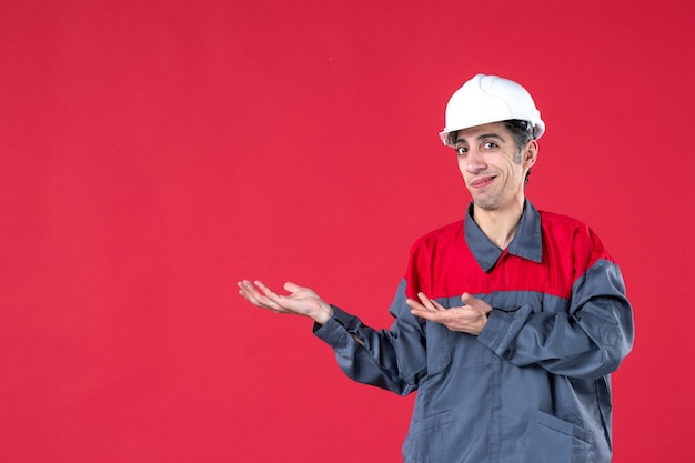 Vista superior del trabajador joven confundido en uniforme con casco apuntando hacia el lado derecho en la pared roja aislada