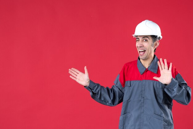 Vista superior del trabajador joven divertido sonriente en uniforme con casco y apuntando algo en el lado derecho que muestra cinco en la pared roja aislada