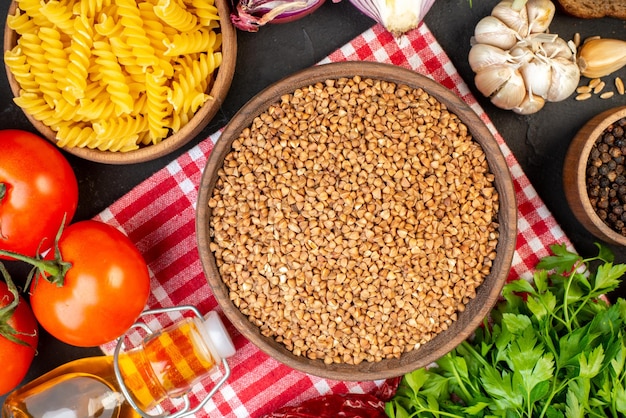 Vista superior de trigo sarraceno crudo en un tazón marrón sobre pasta de toalla en una olla verduras frescas botella de aceite caído sal paquete verde rebanadas de pan