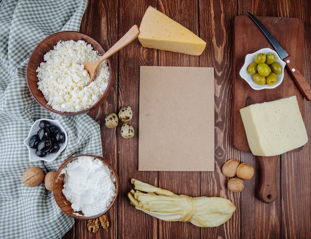 Vista superior de un trozo de hoja de papel marrón y queso cottage en cuencos de madera y varios quesos con aceitunas en escabeche, huevos de codorniz y nueces sobre tela escocesa sobre mesa rústica de madera