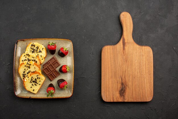 Vista superior de trozos de pastel apetitosos trozos de pastel con chocolate y fresas junto a la tabla de cortar de madera en la mesa oscura