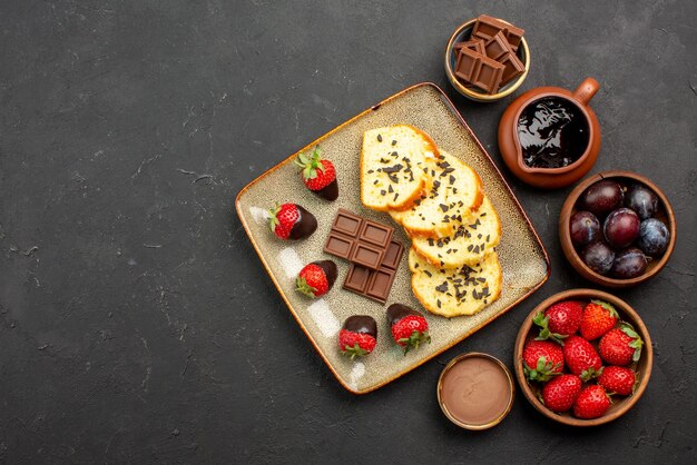 Vista superior de trozos de pastel apetitosos trozos de pastel con chocolate y fresas y tazones con fresas, bayas y salsa de chocolate en el lado derecho de la mesa