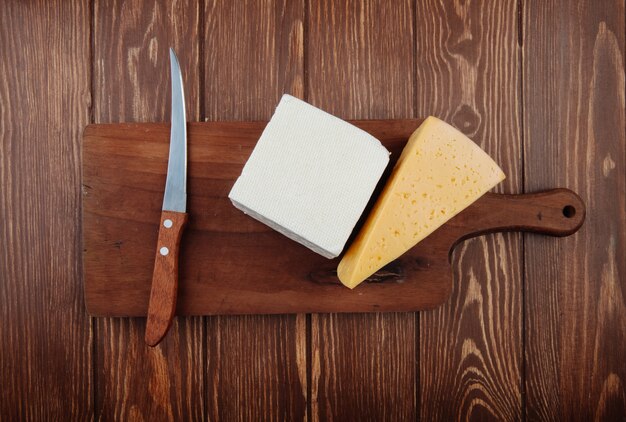 Vista superior de trozos de queso con cuchillo de cocina en una tabla de cortar de madera en la mesa de madera rústica
