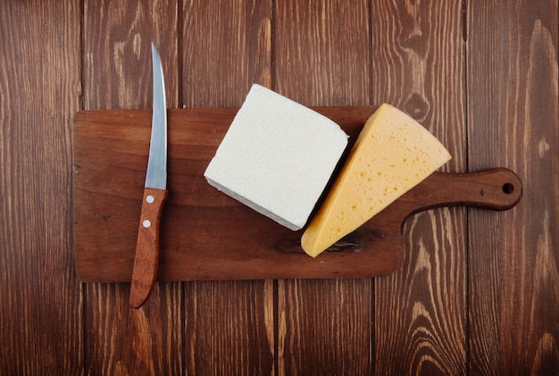 Vista superior de trozos de queso con cuchillo de cocina en una tabla de cortar de madera en la mesa de madera rústica