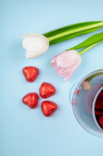 Vista superior de tulipanes de color blanco y rosa con una copa de vino y caramelos de chocolate en forma de corazón en papel rojo esparcidos sobre la mesa azul