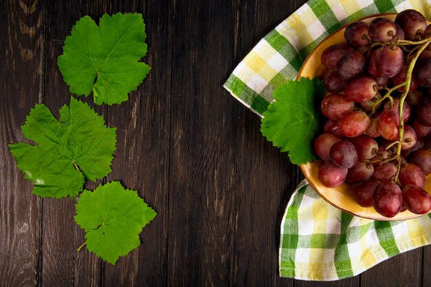 Vista superior de uva dulce fresca en un plato con hojas de uva verde en la mesa de madera oscura con espacio de copia