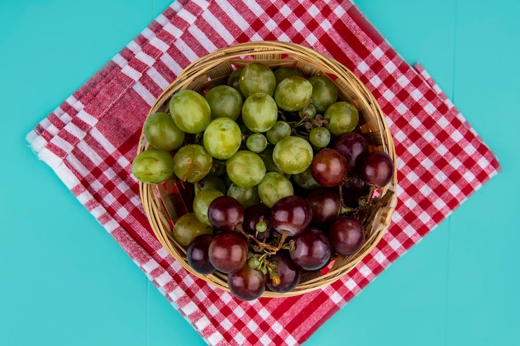 Foto gratuito vista superior de uvas blancas y negras en canasta sobre tela escocesa sobre fondo azul.