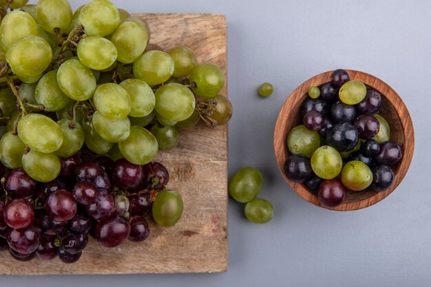 Vista superior de uvas blancas y rojas en tabla de cortar y tazón de uva sobre fondo gris