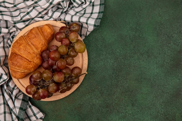 Vista superior de las uvas en una tabla de cocina de madera con croissant sobre un paño marcado sobre una superficie verde con espacio de copia